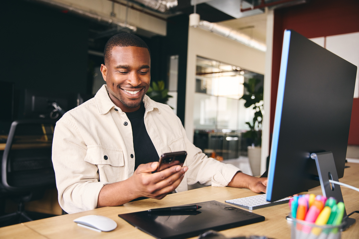 homem segurando celular sentado de frente para um notebook em um escritório